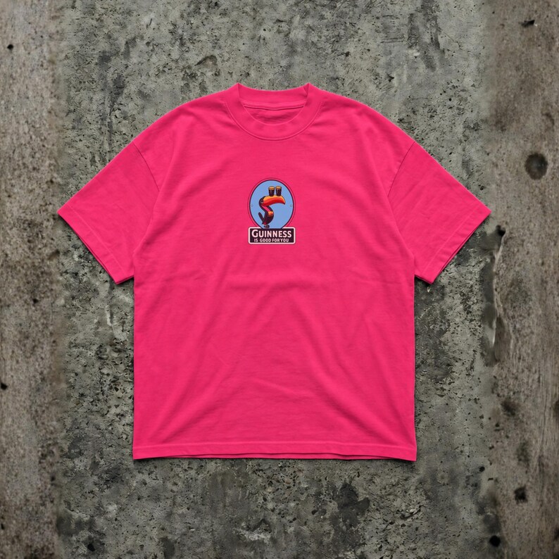 K&ouml;nnte beinhalten: Ein leuchtend pinkfarbenes T-Shirt mit einem Guinness-Logo. Das Logo zeigt einen Tukan in einem blauen Kreis, mit den Worten "Guinness" und "It's good for you" in einem Rechteck. Das T-Shirt hat einen Rundhalsausschnitt und kurze &Auml;rmel.