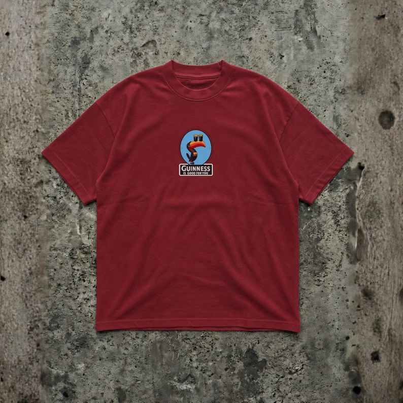 K&ouml;nnte beinhalten: Ein weinrotes T-Shirt mit kurzen &Auml;rmeln und einem kreisf&ouml;rmigen Logo mit einem Tukan, zwei blauen Kreisen und dem Text "Guinness" in einem wei&szlig;en Rechteck. Das Shirt ist aus weichem Material.