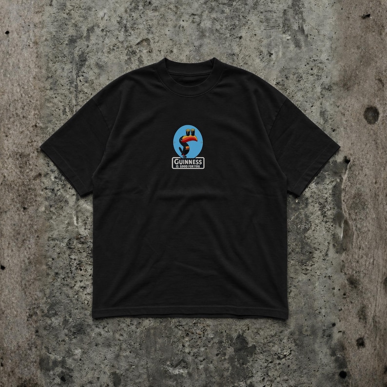 K&ouml;nnte beinhalten: Schwarzes T-Shirt mit einem Guinness-Logo, das einen Tukan zeigt, der zwei Pints Bier h&auml;lt. Das Logo befindet sich vor blauem Hintergrund. Das Guinness-Logo befindet sich unter dem Tukan. Das Shirt ist aus weichem Material.