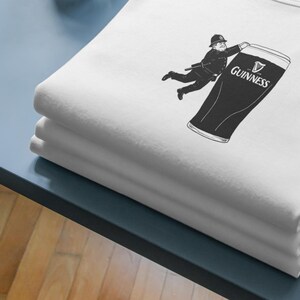 May include: White t-shirt with a black graphic of a police officer reaching for a pint of Guinness. The Guinness logo is visible on the glass. The shirt is folded and stacked on a blue surface.