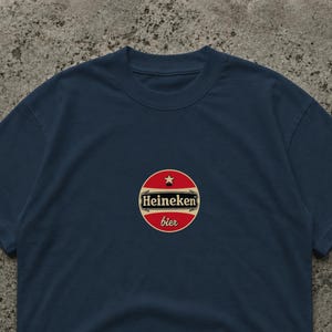 May include: Navy blue t-shirt featuring a circular logo with the text "Heineken bier". The logo is red with a white border and a star at the top. The shirt is a classic crew neck style.