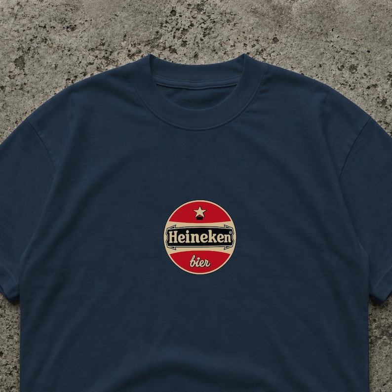 May include: Navy blue t-shirt featuring a circular logo with the text "Heineken bier". The logo is red with a white border and a star at the top. The shirt is a classic crew neck style.