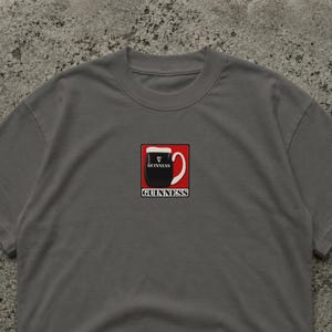 Puede incluir: Camiseta gris con un gr&aacute;fico de una pinta de Guinness en un cuadrado rojo. La pinta es negra con espuma blanca, y la palabra "GUINNESS" est&aacute; impresa arriba y abajo de la pinta en blanco.