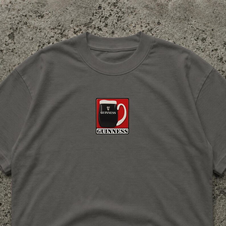 Puede incluir: Camiseta gris con un gr&aacute;fico de una pinta de Guinness en un cuadrado rojo. La pinta es negra con espuma blanca, y la palabra "GUINNESS" est&aacute; impresa arriba y abajo de la pinta en blanco.