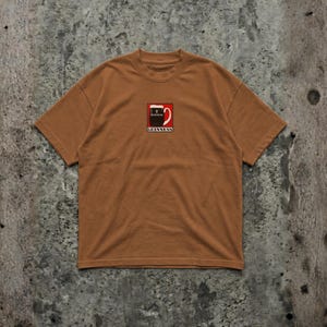May include: A brown t-shirt featuring a Guinness logo on the front. The logo is a stylized pint of Guinness with a red and white design. The shirt has short sleeves and a crew neck.