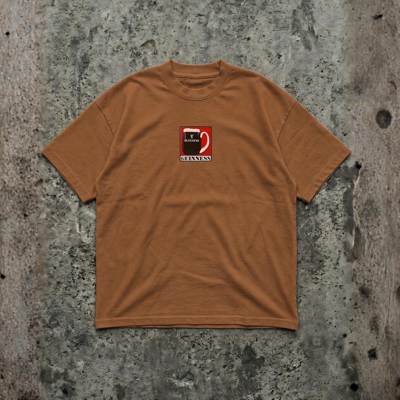 May include: A brown t-shirt featuring a Guinness logo on the front. The logo is a stylized pint of Guinness with a red and white design. The shirt has short sleeves and a crew neck.