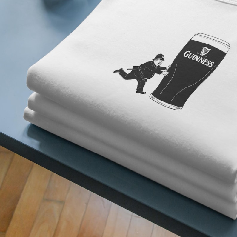 May include: White t-shirt featuring a black and white illustration of a person pushing a large Guinness pint. The Guinness logo is visible on the pint. The t-shirt is folded and stacked.
