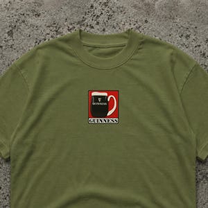 May include: Olive green t-shirt featuring a square graphic of a Guinness pint. The graphic includes a black pint with white foam, a red border, and the word "GUINNESS" in white. The shirt is made of a soft material.