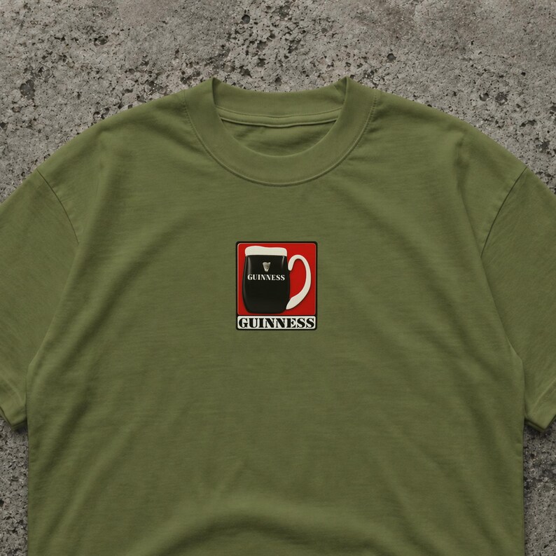 May include: Olive green t-shirt featuring a square graphic of a Guinness pint. The graphic includes a black pint with white foam, a red border, and the word "GUINNESS" in white. The shirt is made of a soft material.
