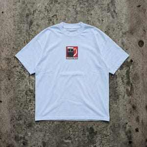 May include: Light blue t-shirt featuring a Guinness logo on the front. The logo is a stylized pint of Guinness with the brand name below. The shirt has a crew neck and short sleeves.