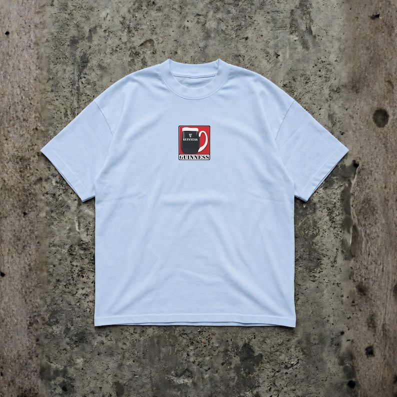 May include: Light blue t-shirt featuring a Guinness logo on the front. The logo is a stylized pint of Guinness with the brand name below. The shirt has a crew neck and short sleeves.