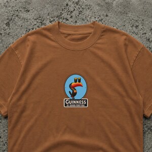 K&ouml;nnte beinhalten: Ein braunes T-Shirt mit einem kreisf&ouml;rmigen Aufdruck, der einen Tukan und zwei Biergl&auml;ser zeigt. Der Aufdruck enth&auml;lt den Text "Guinness is good for you" in einem wei&szlig;en Rechteck. Das Shirt ist aus weichem Material.