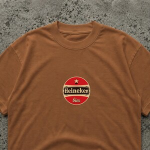 May include: A brown t-shirt featuring a circular logo with the text "Heineken bier" in white and red. The logo includes a star and is centered on the chest of the shirt. The shirt has a crew neck.