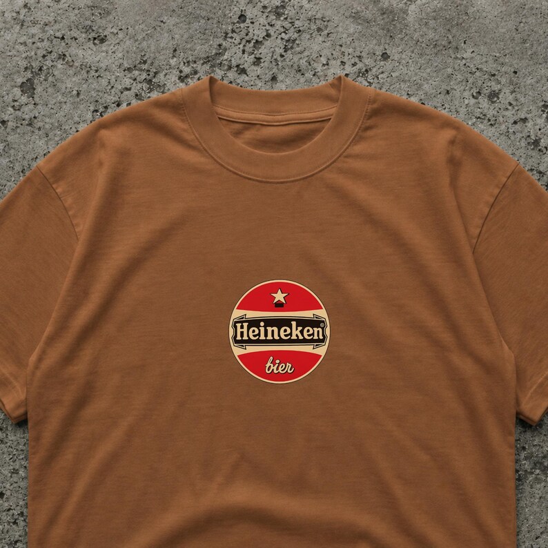 May include: A brown t-shirt featuring a circular logo with the text "Heineken bier" in white and red. The logo includes a star and is centered on the chest of the shirt. The shirt has a crew neck.