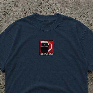 Puede incluir: Camiseta azul marino con un gr&aacute;fico de pinta de Guinness en un cuadrado rojo. La pinta es negra con espuma blanca, y la palabra "Guinness" est&aacute; impresa debajo en blanco. Estilo casual.