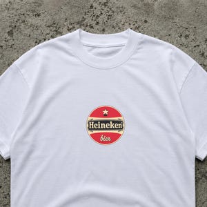 May include: White crew neck t-shirt featuring a circular logo with the text "Heineken bier". The logo is red with a gold border and a small star at the top. The shirt is made of a soft, comfortable material.