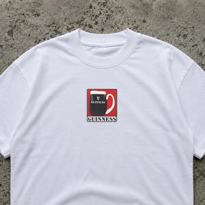 K&ouml;nnte beinhalten: Wei&szlig;es T-Shirt mit einem Guinness-Pint-Motiv. Das Design zeigt ein schwarzes Pint Stout in einem roten Quadrat, mit dem Guinness-Logo oberhalb und unterhalb der Pint-Illustration.