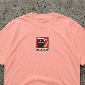 K&ouml;nnte beinhalten: Hellrosa T-Shirt mit einem Guinness-Pint-Motiv. Das Motiv zeigt ein schwarzes Pint Stout mit wei&szlig;em Schaum, einem wei&szlig;en Griff und dem Wort "GUINNESS" in Wei&szlig;. Ein l&auml;ssiges Kleidungsst&uuml;ck.