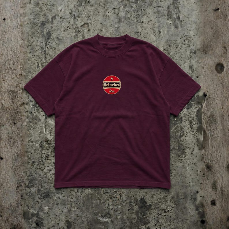 Pu&ograve; includere: T-shirt bordeaux con scollo tondo. La maglietta presenta un logo circolare sul petto con le parole "Heineken" e "bier" in rosso, oro e bianco. La maglietta &egrave; realizzata in materiale morbido.