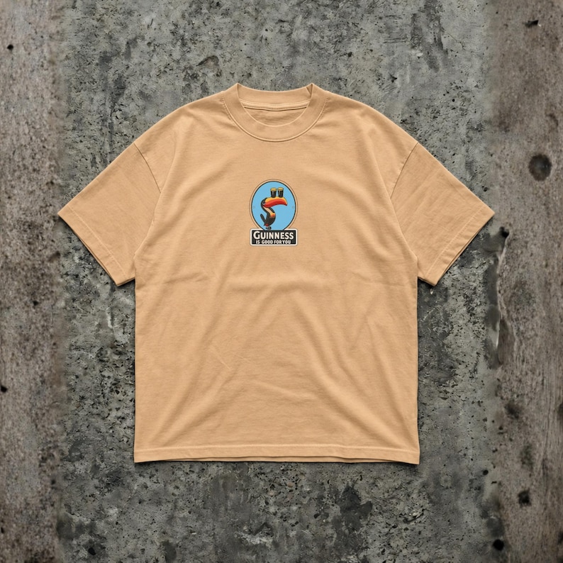 K&ouml;nnte beinhalten: Beigefarbenes T-Shirt mit einem Guinness-Logo auf der Vorderseite. Das Logo zeigt einen Tukan, ein Pint Guinness und den Text "Guinness is good for you". Das T-Shirt hat einen Rundhalsausschnitt und kurze &Auml;rmel.