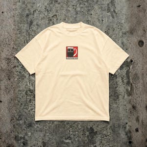 May include: Cream-colored t-shirt featuring a small, centered graphic of a Guinness pint glass. The graphic is in red, black, and white, with the Guinness logo below. The shirt has a classic crew neck and short sleeves.