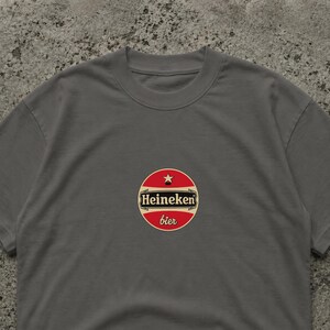 Pu&ograve; includere: Una maglietta grigia con un logo circolare con la scritta "Heineken bier" in rosso e bianco. Il logo include una stella ed &egrave; centrato sul petto. La maglietta &egrave; realizzata in un materiale morbido.
