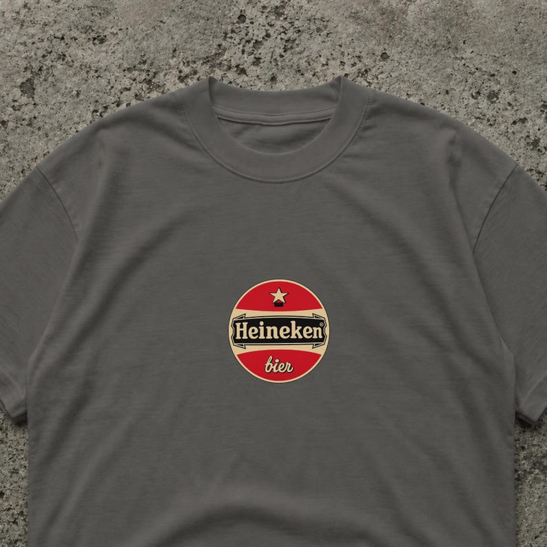 Pu&ograve; includere: Una maglietta grigia con un logo circolare con la scritta "Heineken bier" in rosso e bianco. Il logo include una stella ed &egrave; centrato sul petto. La maglietta &egrave; realizzata in un materiale morbido.