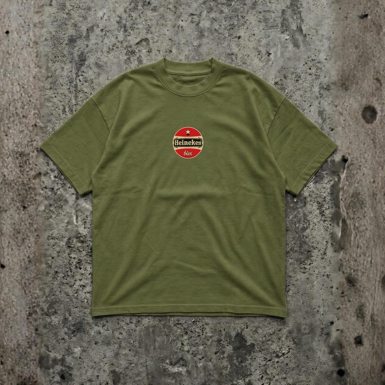 Pu&ograve; includere: T-shirt verde oliva con scollo tondo. La maglietta presenta un logo circolare con una stella rossa e la scritta "Heineken bier". La maglietta &egrave; realizzata in un materiale morbido.
