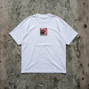 May include: White t-shirt featuring a Guinness logo. The logo is a stylized pint of Guinness with a red background and the word "Guinness" below. The shirt has short sleeves and a crew neck.