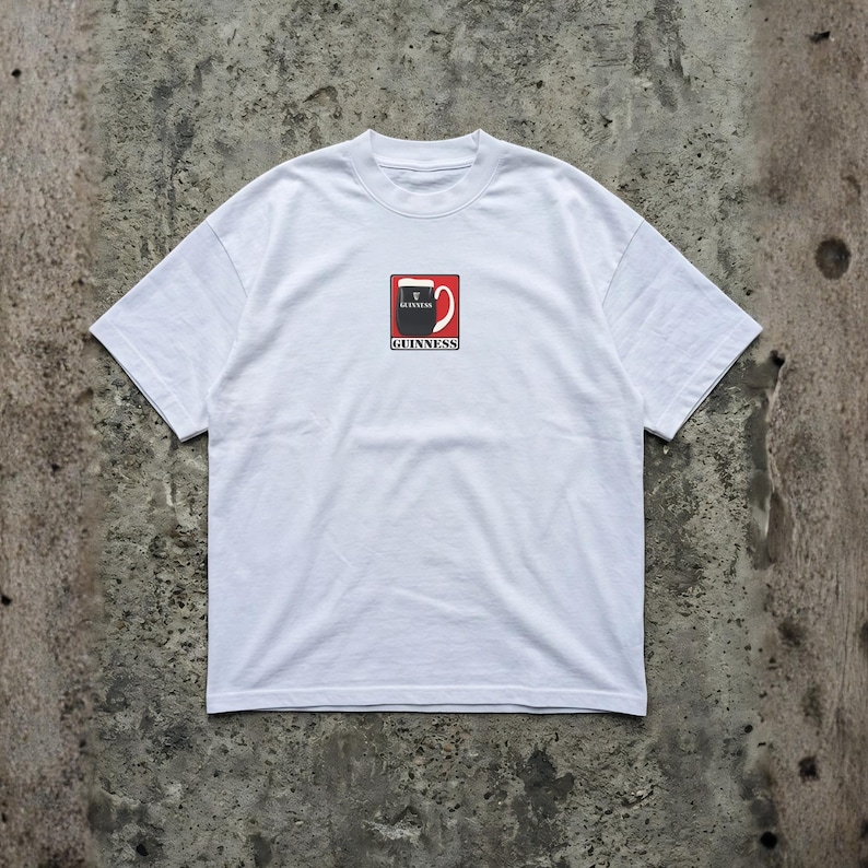 May include: White t-shirt featuring a Guinness logo. The logo is a stylized pint of Guinness with a red background and the word "Guinness" below. The shirt has short sleeves and a crew neck.
