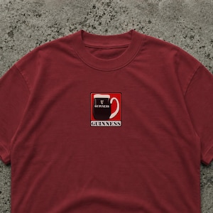 May include: A maroon t-shirt featuring a graphic of a pint of Guinness stout in a square frame. The pint is black with a white head, and the word "Guinness" is printed above and below the image in white.