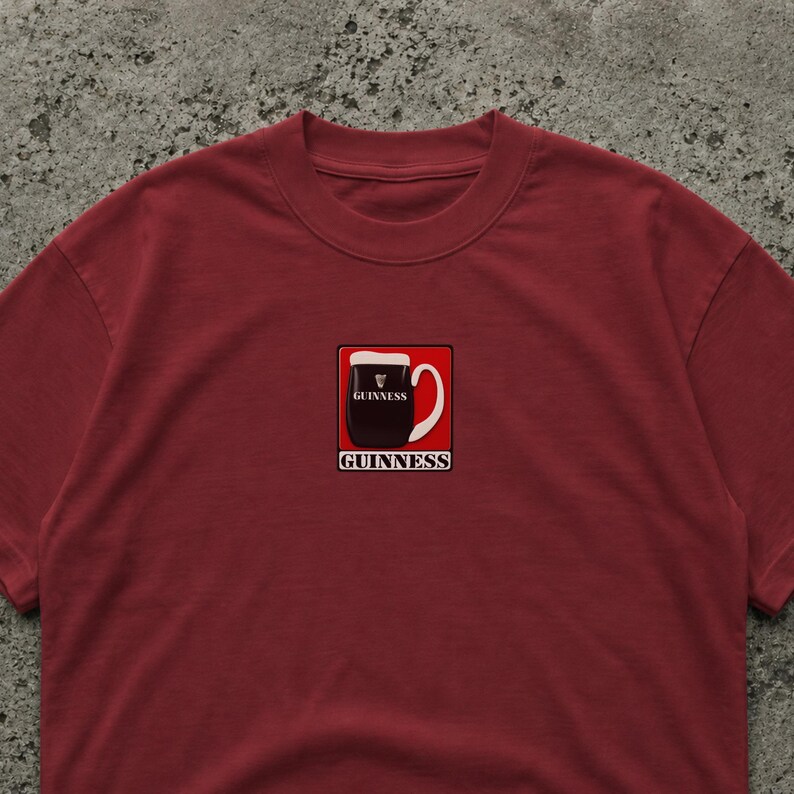 May include: A maroon t-shirt featuring a graphic of a pint of Guinness stout in a square frame. The pint is black with a white head, and the word "Guinness" is printed above and below the image in white.