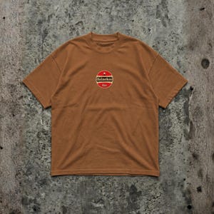 Pu&ograve; includere: T-shirt marrone con girocollo e maniche corte. Presenta un logo circolare con una stella rossa, una striscia bianca e le parole "Heineken" e "bier" in un font stilizzato. La maglietta &egrave; realizzata in materiale morbido.