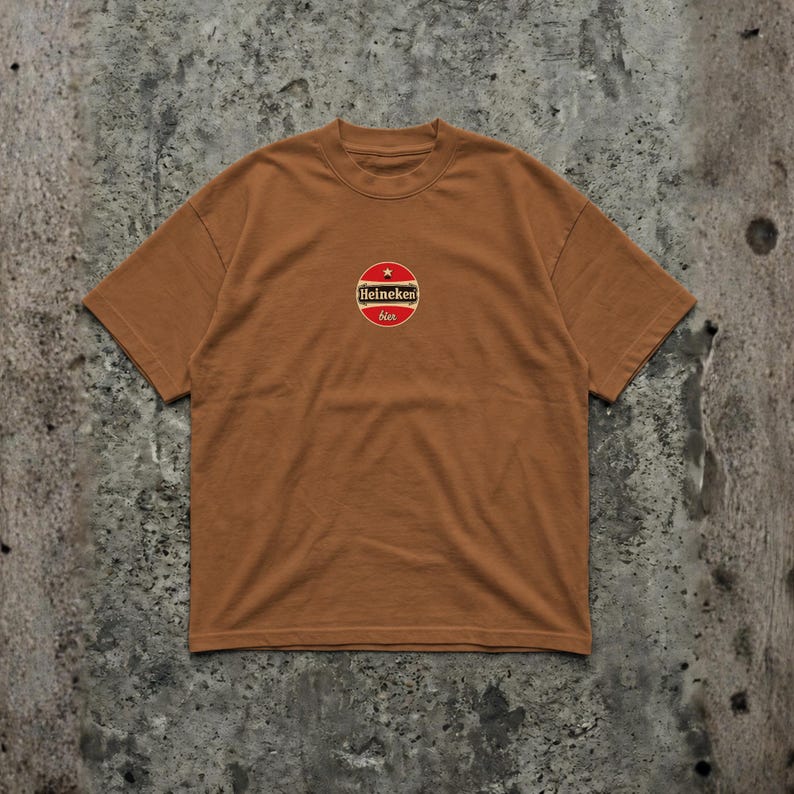 Pu&ograve; includere: T-shirt marrone con girocollo e maniche corte. Presenta un logo circolare con una stella rossa, una striscia bianca e le parole "Heineken" e "bier" in un font stilizzato. La maglietta &egrave; realizzata in materiale morbido.