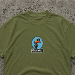 May include: Olive green t-shirt featuring a colorful graphic of a toucan with two pints of Guinness. The toucan is inside a blue circle. Below the graphic is the text "Guinness IS GOOD FOR YOU" in a white rectangle.