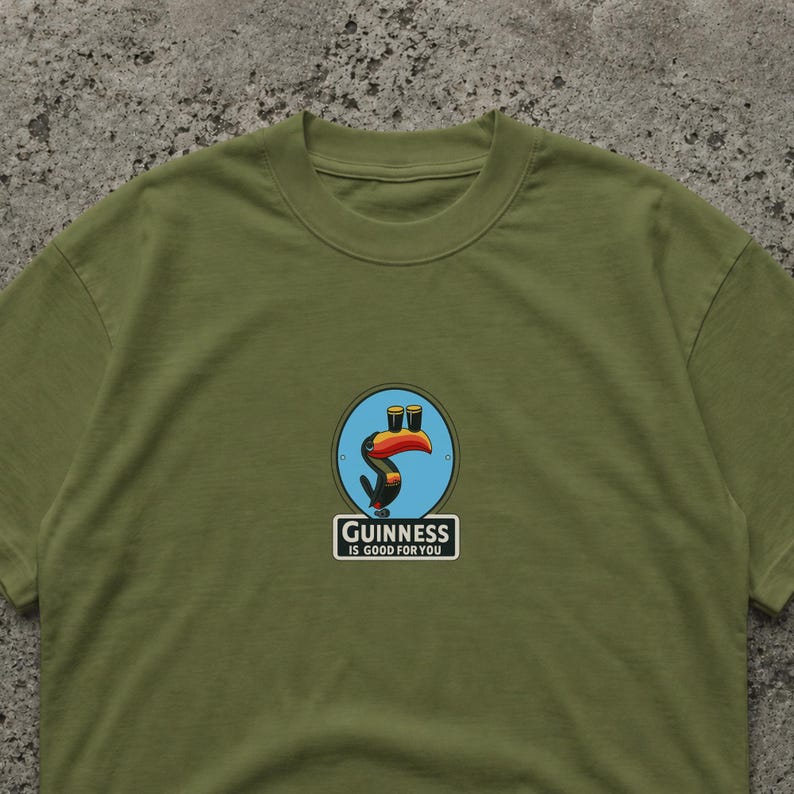 May include: Olive green t-shirt featuring a colorful graphic of a toucan with two pints of Guinness. The toucan is inside a blue circle. Below the graphic is the text "Guinness IS GOOD FOR YOU" in a white rectangle.