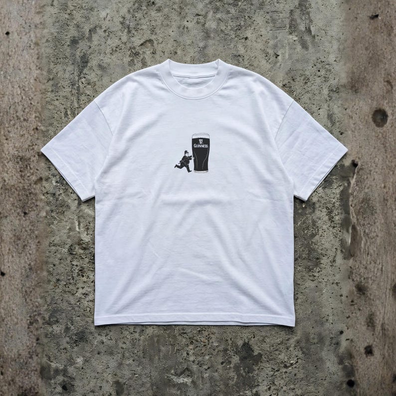 May include: White t-shirt featuring a black graphic of a pint of Guinness with a running figure. The Guinness logo is visible at the top of the pint. The shirt is made of a soft, comfortable material.