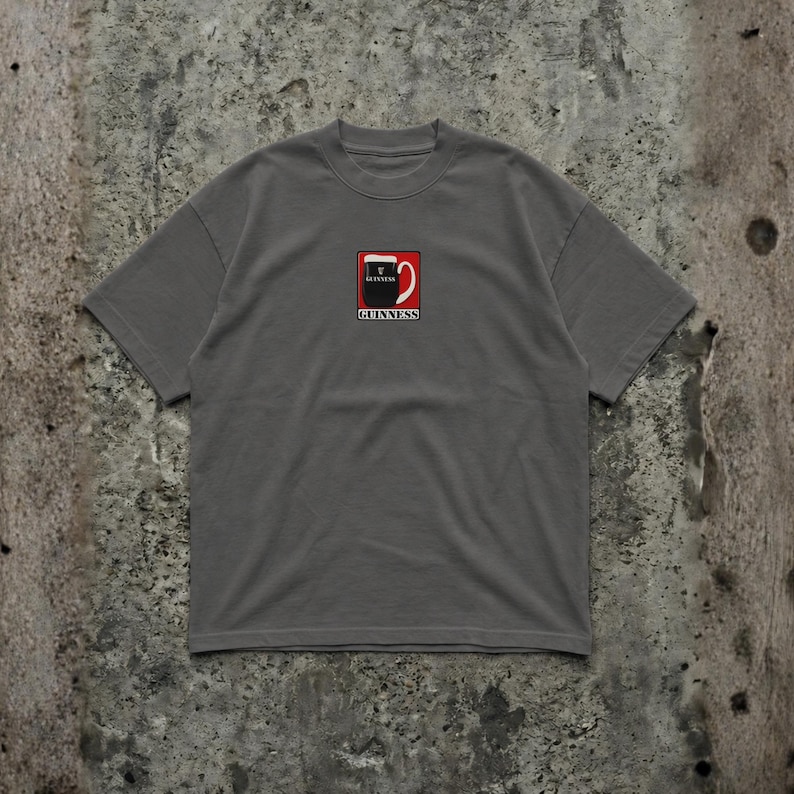 May include: A gray t-shirt featuring a small embroidered design of a Guinness pint glass. The design includes the Guinness logo in red, black, and white. The shirt has short sleeves and a crew neck.