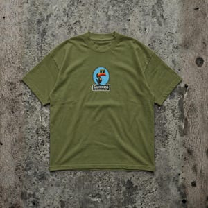 K&ouml;nnte beinhalten: Olivgr&uuml;nes T-Shirt mit einem kreisf&ouml;rmigen Logo, das einen Tukan und den Text "Guinness is good for you" zeigt. Das Logo hat einen roten Schnabel. Das T-Shirt hat einen Rundhalsausschnitt und kurze &Auml;rmel.