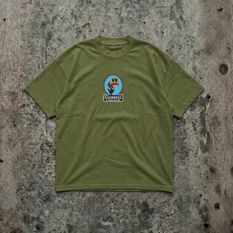 K&ouml;nnte beinhalten: Olivgr&uuml;nes T-Shirt mit einem kreisf&ouml;rmigen Logo, das einen Tukan und den Text "Guinness is good for you" zeigt. Das Logo hat einen roten Schnabel. Das T-Shirt hat einen Rundhalsausschnitt und kurze &Auml;rmel.