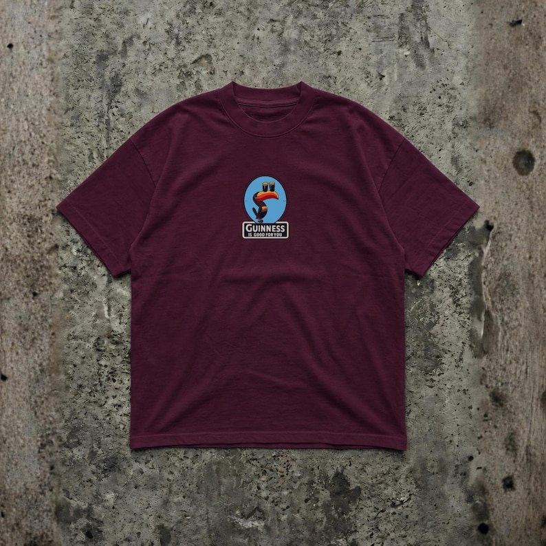 K&ouml;nnte beinhalten: Burgunderfarbenes T-Shirt mit einem Guinness-Logo, das einen Tukan mit einem Guinness-Pint zeigt, mit dem Text "Guinness is good for you". Das Shirt hat einen Rundhalsausschnitt und kurze &Auml;rmel.