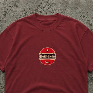 Op de afbeelding: Een bordeauxrood T-shirt met een cirkelvormig logo met de tekst "Heineken bier". Het logo is rood, wit en zwart met een ster. Het T-shirt is effen en heeft een ronde hals.