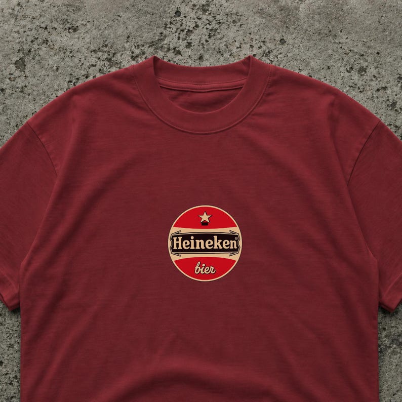 Op de afbeelding: Een bordeauxrood T-shirt met een cirkelvormig logo met de tekst "Heineken bier". Het logo is rood, wit en zwart met een ster. Het T-shirt is effen en heeft een ronde hals.
