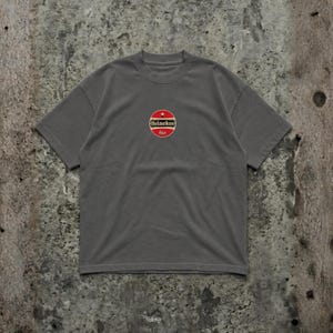 Pu&ograve; includere: T-shirt grigio con logo circolare con la scritta "Heineken bier" in rosso e bianco. Il logo &egrave; centrato sul petto. La maglietta ha maniche corte e girocollo.
