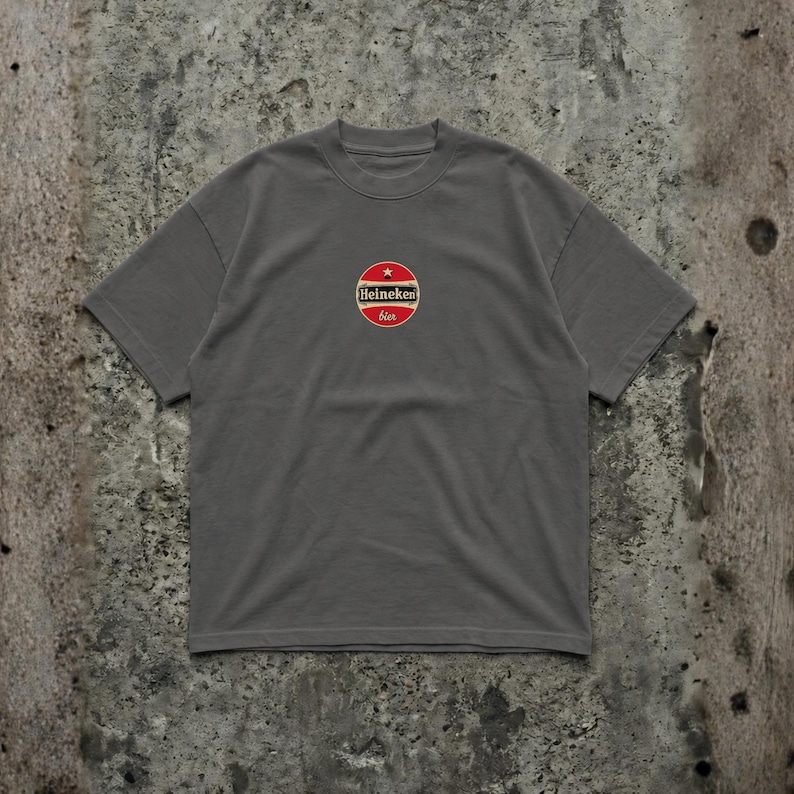Pu&ograve; includere: T-shirt grigio con logo circolare con la scritta "Heineken bier" in rosso e bianco. Il logo &egrave; centrato sul petto. La maglietta ha maniche corte e girocollo.