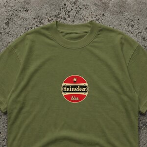 May include: Olive green t-shirt featuring a circular logo with the text "Heineken bier". The logo is red with a white border and a star at the top. The shirt is made of a soft material.