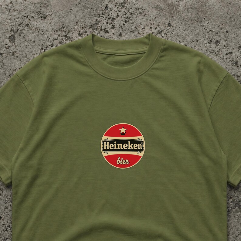 May include: Olive green t-shirt featuring a circular logo with the text "Heineken bier". The logo is red with a white border and a star at the top. The shirt is made of a soft material.