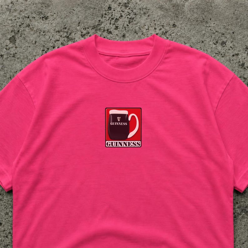 K&ouml;nnte beinhalten: Ein leuchtend pinkfarbenes T-Shirt mit einem Guinness-Pint-Motiv. Das Motiv zeigt ein schwarzes Pint mit wei&szlig;em Schaum und einem wei&szlig;en Griff, eingebettet in ein rotes Quadrat. Das Wort "GUINNESS" steht dar&uuml;ber und darunter.