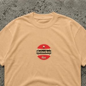 Puede incluir: Camiseta color beige con cuello redondo y un dise&ntilde;o circular. El dise&ntilde;o tiene un fondo rojo con una estrella blanca y la palabra "Heineken" en negro. Debajo est&aacute; la palabra "bier" en rojo.