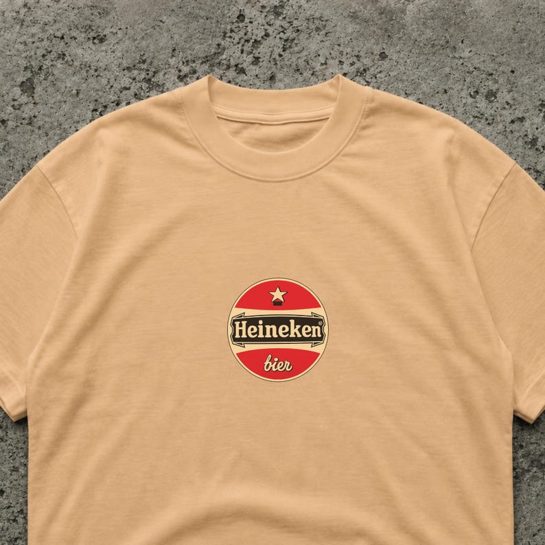 Puede incluir: Camiseta color beige con cuello redondo y un dise&ntilde;o circular. El dise&ntilde;o tiene un fondo rojo con una estrella blanca y la palabra "Heineken" en negro. Debajo est&aacute; la palabra "bier" en rojo.